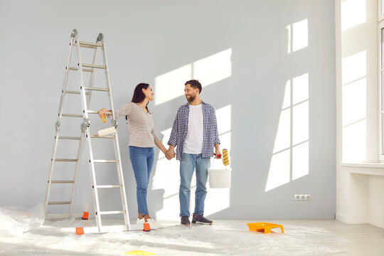 Happy Young Couple Holding Hands While Painting Walls In Their New Home. Cheerful Husband And Wife With Paint Rollers Renovating And Decorating Room In Their New Home