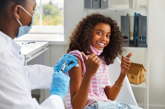 Childhood immunization. HHappy dark-skinned teenage girl showing thumbs up gesture while being immunized against viral infections. Pediatrician makes vaccination to cute schoolgirl in clinic. - Powered by Adobe