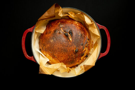 Fresh sourdough bread loaf in parchment paper inside a red enamel cast iron pot against a black background