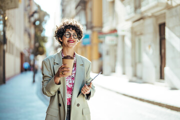 Obraz premium Dreamy woman in formal clothes carries notepad tablet disposable cup of coffee has walk outdoor during daytime poses near office building with cheerful expression going to have job interview