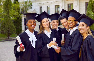 Happy graduate students posing for photo in front of university building. Successful classmates in black mortar board caps and bachelor gowns with diplomas celebrating college or university graduation