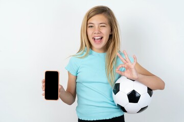 beautiful caucasian teen girl wearing sportswear holding a football ball over white wall holding in...