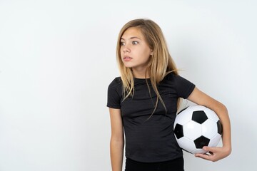 beautiful caucasian teen girl wearing sportswear holding a football ball over white wall stares aside with wondered expression has speechless expression. Embarrassed model looks in surprise