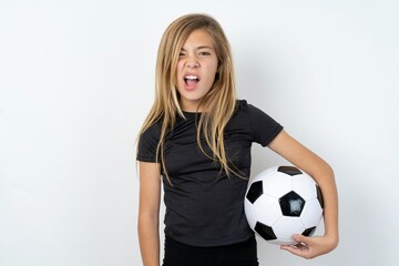 Portrait of dissatisfied beautiful caucasian teen girl wearing sportswear holding a football ball, smirks face, purses lips and looks with annoyance at camera, discontent hearing something unpleasant