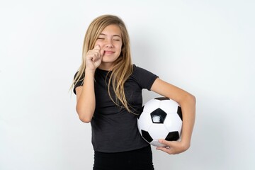 Pleased beautiful caucasian teen girl wearing sportswear holding a football ball over white wall with closed eyes keeps hands near cheeks and smiles tenderly imagines something very pleasant