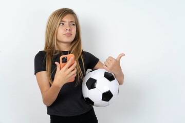 beautiful caucasian teen girl wearing sportswear  over white wall points thumb away and shows blank space aside, holds mobile phone for sending text messages.