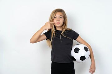 Charming beautiful caucasian teen girl wearing sportswear and football ball over white wall pointing on pout lips with forefinger, showing effect after lifting procedure,