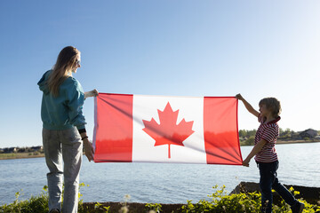 Fototapeta woman and a child, mother and son, hold the Canadian flag on a sunny day against the background of the sky and the river obraz