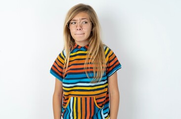 Amazed puzzled beautiful caucasian teen girl wearing striped T-shirt over white wall, curves lips and has worried look, sees something awful in front.