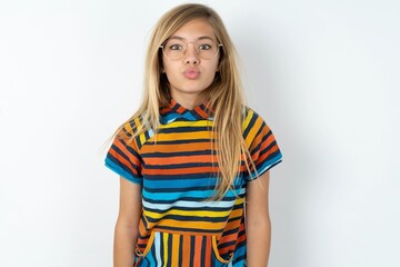 beautiful caucasian teen girl wearing striped T-shirt over white wall, keeps lips as going to kiss someone, has glad expression, grimace face. Standing indoors. Beauty concept.