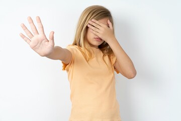 beautiful caucasian teen girl wearing orange T-shirt over white wall covers eyes with palm and...