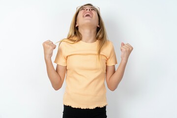 beautiful caucasian teen girl wearing orange T-shirt over white wall looks with excitement up, keeps hands raised, notices something unexpected.