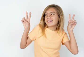 Isolated shot of cheerful beautiful caucasian teen girl wearing orange T-shirt over white wall makes peace or victory sign with both hands, feels cool.