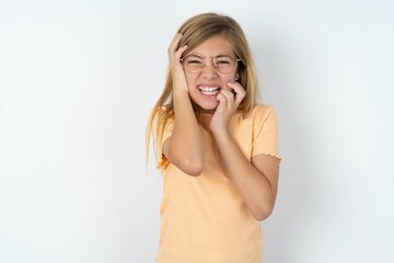 Young gloomy beautiful caucasian teen girl wearing orange T-shirt over white wall, hiding face with...