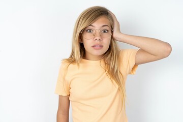 Portrait of confused beautiful caucasian teen girl wearing orange T-shirt over white wall holding hand on hair and frowning, panicking, losing memory. Worried and anxious can not remember anything.