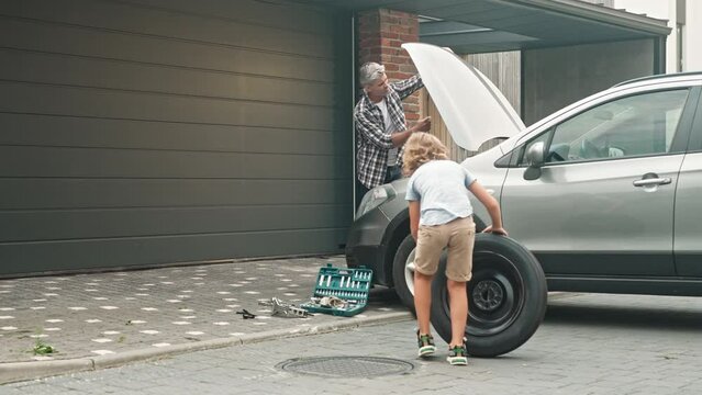 Portrait of adult man lifting hood of auto on background of home garage. Pre-adolescent cute boy in yard swings spare tire to car. Dad and son work as team and repair vehicle.