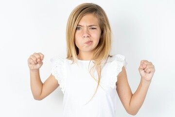 Irritated beautiful caucasian teen girl wearing white T-shirt over white wall blows cheeks with anger and raises clenched fists expresses rage and aggressive emotions. Furious model