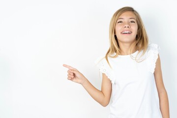 beautiful caucasian teen girl wearing white T-shirt over white wall laughs happily points away on blank space demonstrates shopping discount offer, excited by good news or unexpected sale.