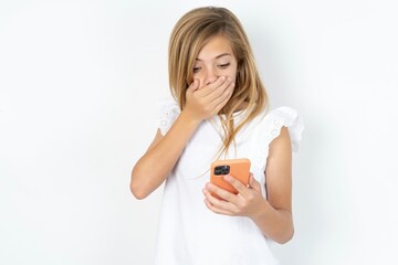 beautiful caucasian teen girl wearing white T-shirt over white wall being deeply surprised, stares at smartphone display, reads shocking news on website, Omg, its horrible!