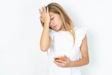 Upset depressed beautiful caucasian teen girl wearing white T-shirt over white wall makes face palm as forgot about something important holds mobile phone expresses sorrow and regret blames