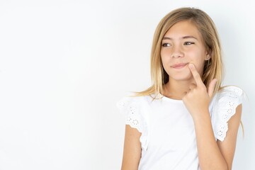 Lovely dreamy beautiful caucasian teen girl wearing white T-shirt over white wall keeps finger near lips looks aside copy space.