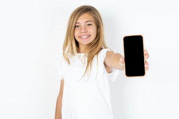 Naklejka premium Charming adorable beautiful caucasian teen girl wearing white T-shirt over white wall holding modern device, showing black screen smartphone