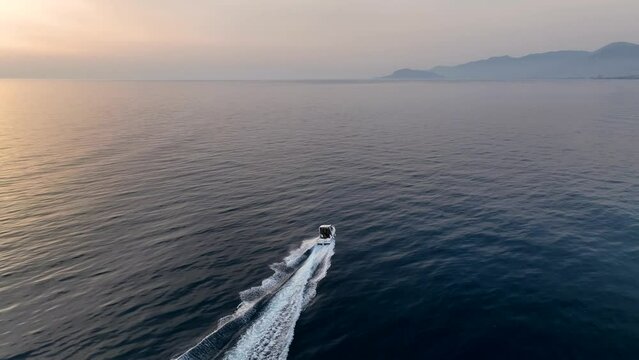 A bird's-eye view of a small speedboat in the bay during sunset. A picture from a drone rushing towards an exotic picturesque seascape. Top view of water sports.