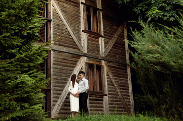 Fototapeta premium beautiful pregnant couple on the background of a wooden ecological house in the forest. a man hugs a pregnant woman.