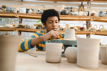 Young African American boy and girl wearing apron preparing pots and sculptures using mud and and clay and painting them in a workshop studio while decorating them using brushes