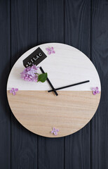On a gray wooden background lies a clock on which the hands point to the inscription lilac