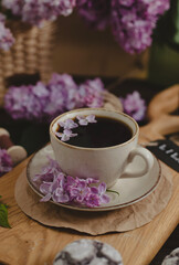 A cup of Americano black coffee, purple lilac flowers on a wooden background. A cup of Americano black coffee, purple lilac flowers on a wooden background. Lilac inscription on the card next to it
