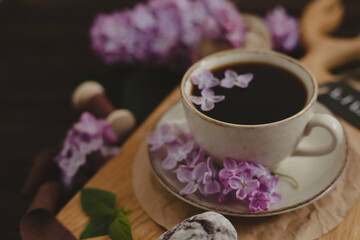 A cup of Americano black coffee, purple lilac flowers on a wooden background. A cup of Americano black coffee, purple lilac flowers on a wooden background. Lilac inscription on the card next to it