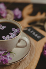 Obraz na płótnie Canvas A cup of Americano black coffee, purple lilac flowers on a wooden background. A cup of Americano black coffee, purple lilac flowers on a wooden background. Lilac inscription on the card next to it