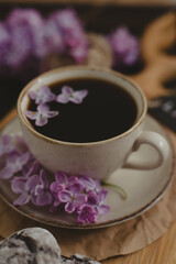 A cup of Americano black coffee, purple lilac flowers on a wooden background. A cup of Americano black coffee, purple lilac flowers on a wooden background. Lilac inscription on the card next to it