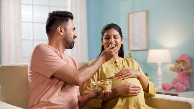 Happy indian husband feeding fruit salad to pregnant wife at home - concept of family caring, relationship bonding and maternal wellness