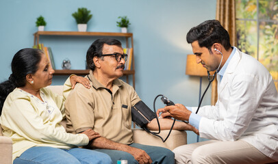 Indian doctor checking bp or blood pressure to senior man while sitting on sofa with wife at home -...