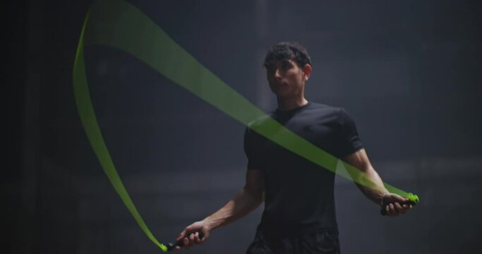 Young handsome Caucasian man boxer jump rope exercises in abandoned building hall gym under smoke light. Skipping man working out in cross training space in slow motion. Silhouette on dark background