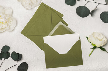 Feminine invitation or greeting card mockup with green envelopes and white peony flowers and eucalyptus sprigs on light background.