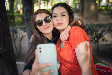 Two young latin female friends walking at the woods. Embrace each other and laughing. Friends making selfie. Two beautiful latin young women making selfie