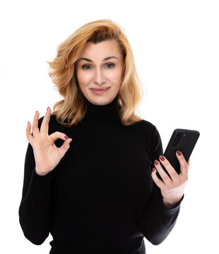 Portrait of forty year old woman shows something interesting on the smartphone screen and make ok okay gesture, isolated on white background. Woman in black turtleneck posing in studio.
