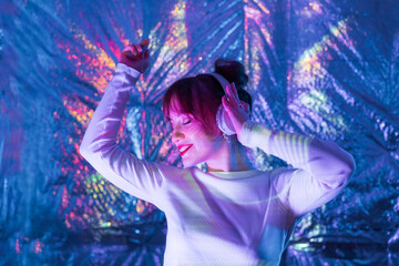 Relaxed smiling woman with closed eyes in white clothes and headphones dancing in neon light. Music lover. White dress code party. Silent disco. Enjoy moment on shiny background. Selective focus.