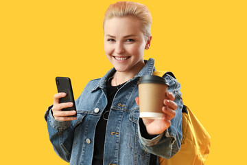 Female student with cup of coffee and mobile phone on yellow background, closeup
