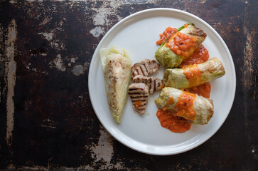 Cabbage rolls with grilled pork tenderloin, cooked cabbage and tomato sauce on a white plate and table, top view