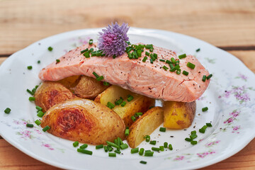 Close up picture on steamed slamon fillet with baked new not pelled potatoes and sprinkled with choped chives and decorated with chives blossom served on rustic style prcelain plate on wooden table.