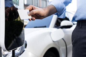 Hand holding credit card to pay public charging station and recharge her electric vehicle,...