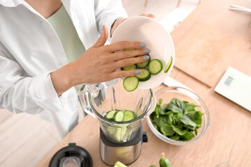 Young woman putting cucumber slices into blender in kitchen, closeup