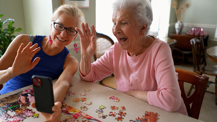 Happy waving and smiling, laughing elderly senior woman sitting at table with mature middle aged daughter, video chat on phone. Smiling talking to family on smartphone.