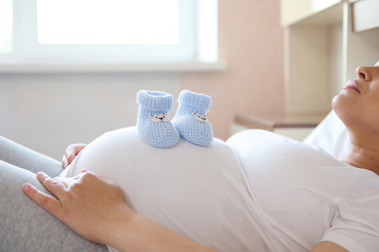 Pregnant woman expecting her future baby, relaxing at home, putting baby booties on her big belly. Pregnancy trimester third, maternity, happy motherhood concept. Close-up advertising shot. Copy space