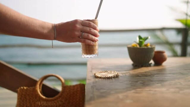 Female hand taking tall glass with cocktail and metal straw from the wooden table in outdoor tropical sea view cafe. Removing iced coffee or smoothie onto coaster in rooftop restaurant with ocean view