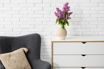 Vase with beautiful lilac flowers on chest of drawers near light brick wall